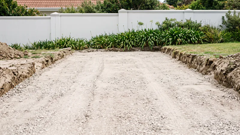 An excavated driveway filled with a levelled layer of pale crushed-stone base material between cut soil banks, in front of a white boundary wall