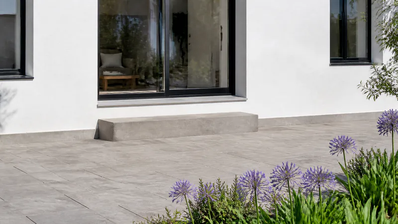 A patio of grey flagstones below a black-framed sliding door, with a single wide concrete step down from the door to the patio and a band of white wall between the paving and the door