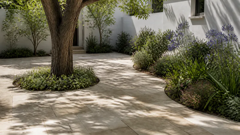 A pale sandstone flagstone patio in dappled shade, with an unpaved planted ring around the trunk of a large tree and a curved bed of agapanthus cut into its edge