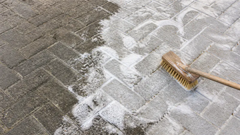 A stiff wooden scrubbing brush on grey herringbone paving, with soapy foam over the scrubbed half, which is visibly lighter than the grimy pavers beside it