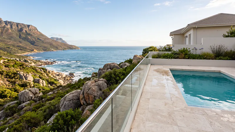 A pale stone pool terrace with a glass balustrade on a hillside above granite boulders, fynbos and the Atlantic Ocean, with mountain cliffs behind